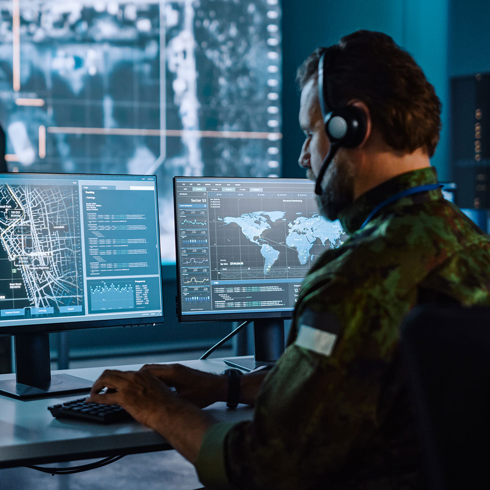 Military operative monitoring digital maps and data on dual screens in a secure command center.